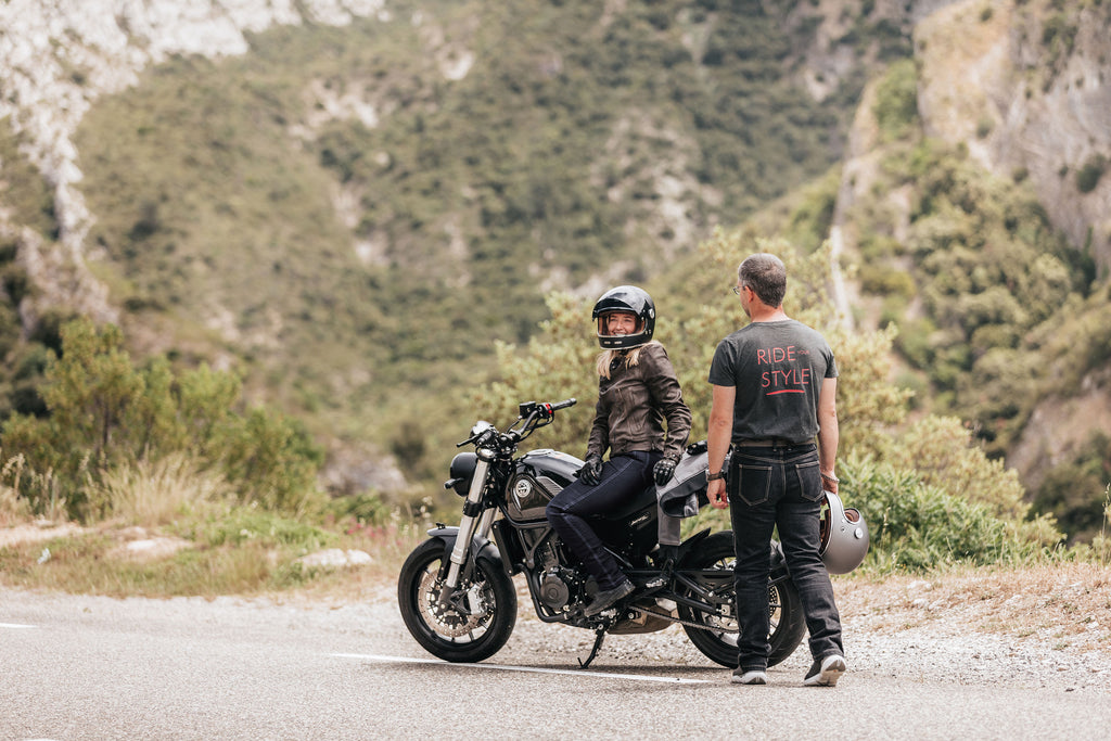 Femme sur une moto portant la veste de motarde bonnette et un homme portant un jean moto noir sur le bord d'une route